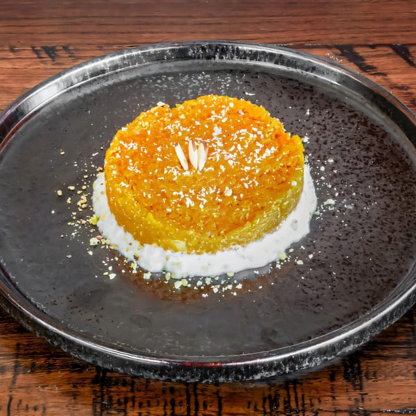 A vibrant orange Indian dessert, garnished with nuts and white flakes, on a dark plate.