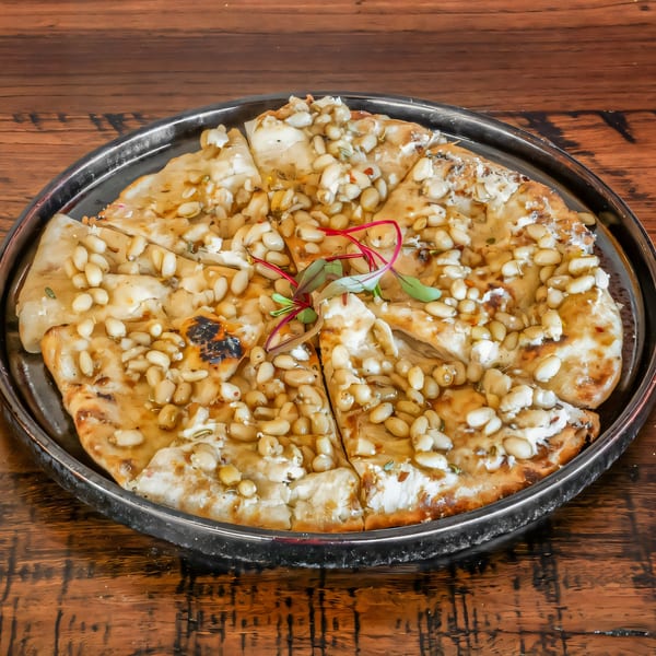Sliced Indian flatbread topped with pine nuts, cream cheese, and microgreens on a dark metal plate.