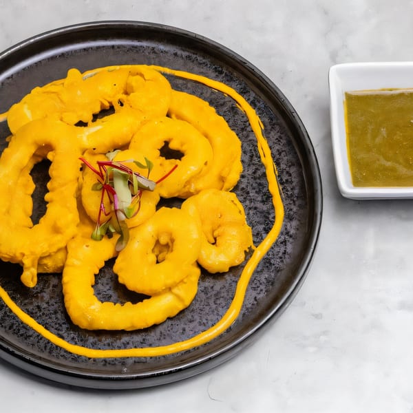 Bright yellow calamari rings with microgreens on a black plate, next to a white bowl of green sauce.